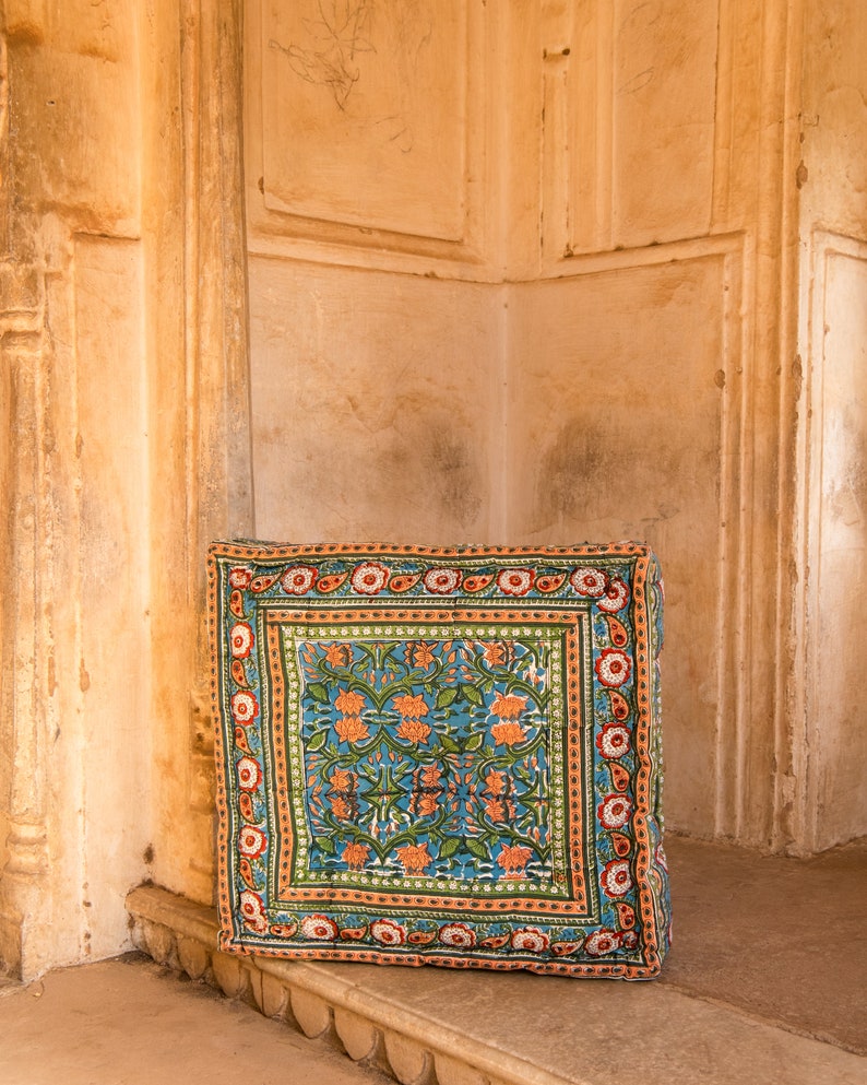 May include: A square floor cushion with a blue, green, orange, and red floral pattern. The cushion is sitting on a stone step.