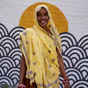 Peut inclure: Un foulard jaune et un ch&acirc;le assorti orn&eacute;s de petits motifs floraux brod&eacute;s en rose, vert et bleu, port&eacute;s sur une jupe marron. Le textile pr&eacute;sente un grand motif de soleil orange au-dessus de vagues r&eacute;p&eacute;titives noires et blanches.