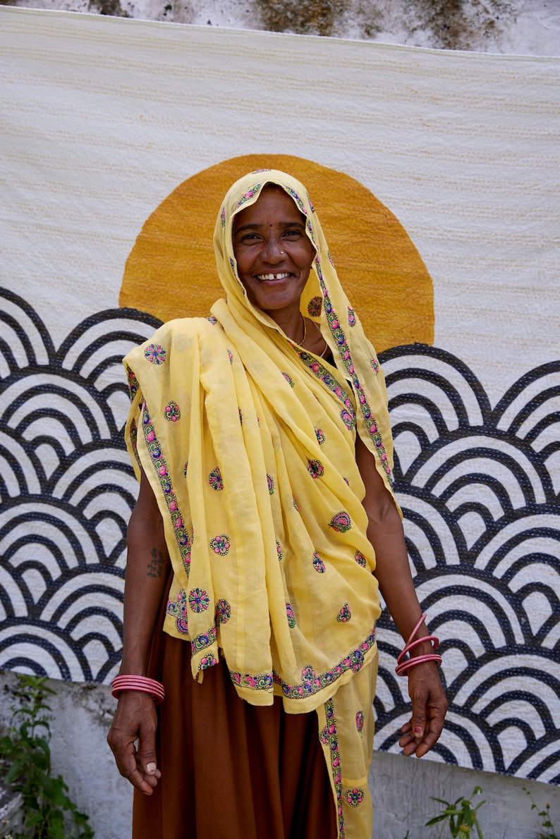Peut inclure: Un foulard jaune et un ch&acirc;le assorti orn&eacute;s de petits motifs floraux brod&eacute;s en rose, vert et bleu, port&eacute;s sur une jupe marron. Le textile pr&eacute;sente un grand motif de soleil orange au-dessus de vagues r&eacute;p&eacute;titives noires et blanches.