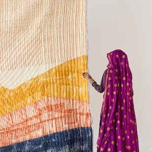 May include: A large, colorful quilt with a geometric design. The quilt features a cream-colored top with vertical orange stripes, transitioning to yellow, orange, and navy blue sections. The quilt is bordered with a striped fabric.