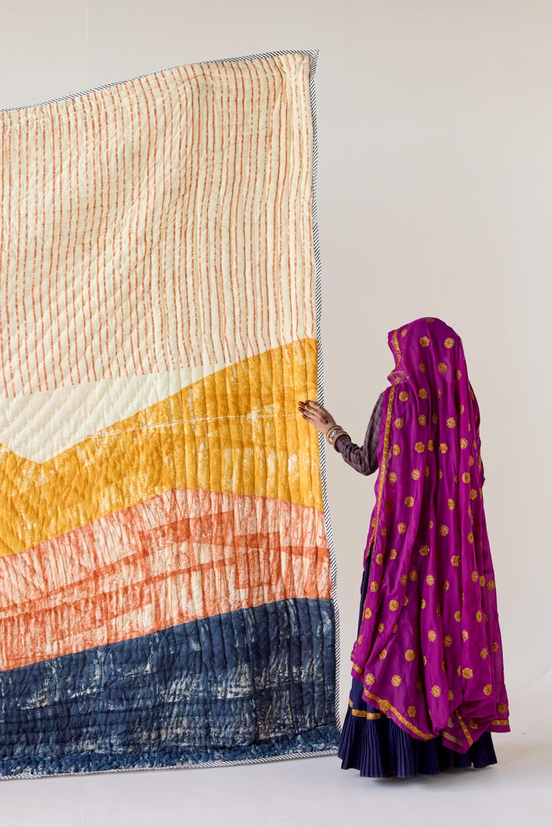 May include: A large, colorful quilt with a geometric design. The quilt features a cream-colored top with vertical orange stripes, transitioning to yellow, orange, and navy blue sections. The quilt is bordered with a striped fabric.