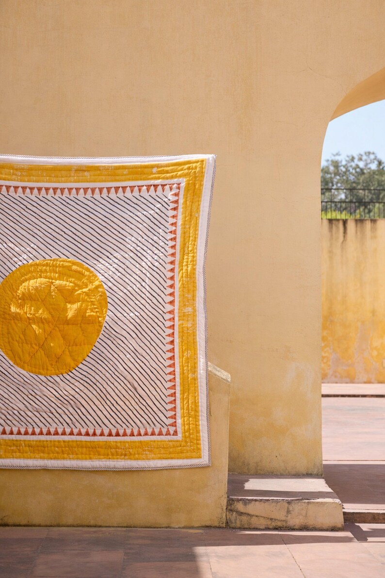 May include: A yellow and white quilt with a large yellow circle in the center. The quilt has black diagonal stripes and a yellow border with orange triangles. The quilt is hanging on a yellow wall.