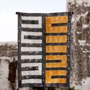 May include: A black and white geometric patterned throw blanket with a contrasting yellow geometric pattern on the right side. The blanket is hanging on a wall.