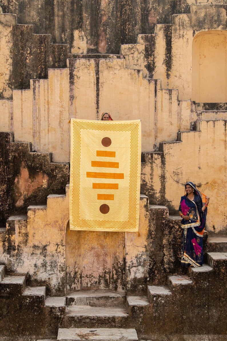 Puede incluir: Dos mujeres se paran frente a una tela amarilla y naranja con un patr&oacute;n que cuelga de una pared de piedra. La tela tiene un dise&ntilde;o geom&eacute;trico con c&iacute;rculos y rect&aacute;ngulos. Las mujeres visten ropa tradicional india.
