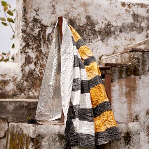 May include: A white and black striped kantha quilt with yellow accents is draped over a wooden chair. The quilt is hanging against a whitewashed wall with a rustic texture.