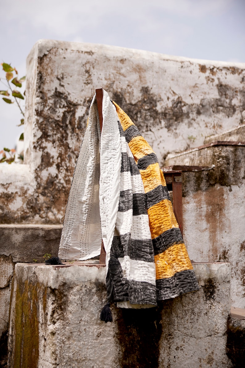 May include: A white and black striped kantha quilt with yellow accents is draped over a wooden chair. The quilt is hanging against a whitewashed wall with a rustic texture.
