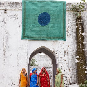 K&ouml;nnte beinhalten: Vier Frauen in farbenfroher traditioneller indischer Kleidung stehen vor einem gew&ouml;lbten Torbogen. &Uuml;ber ihnen h&auml;ngt eine t&uuml;rkisblaue Steppdecke. Die Frauen tragen leuchtende Saris in Gelb, Blau und Rot mit passenden Kopfbedeckungen. Der Hintergrund ist eine wei&szlig;e, verwitterte Wand.