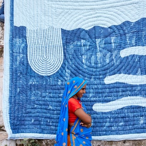 K&ouml;nnte beinhalten: Eine gro&szlig;e, blau-wei&szlig;e Steppdecke mit Landschaftsdesign h&auml;ngt an einer Wand. Die Steppdecke zeigt einen hellblauen Himmel und eine dunklere blaue Landschaft mit geschwungenen Linien und wolkenartigen Formen. Eine Frau in einem blauen Sari steht vor der Steppdecke.