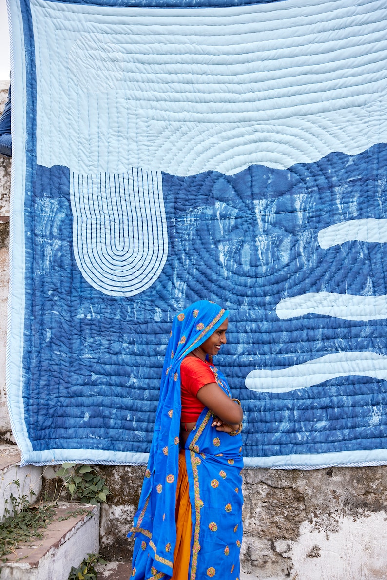 K&ouml;nnte beinhalten: Eine gro&szlig;e, blau-wei&szlig;e Steppdecke mit Landschaftsdesign h&auml;ngt an einer Wand. Die Steppdecke zeigt einen hellblauen Himmel und eine dunklere blaue Landschaft mit geschwungenen Linien und wolkenartigen Formen. Eine Frau in einem blauen Sari steht vor der Steppdecke.