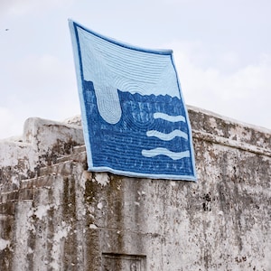 K&ouml;nnte beinhalten: Ein blau-wei&szlig;es Quilt mit einem vom Wasser inspirierten Design. Das Quilt hat einen hellblauen Hintergrund mit dunkelblauen Wellen- und Wasserfallmustern. Das Quilt wird im Freien &uuml;ber einer verwitterten Steinstruktur drapiert.