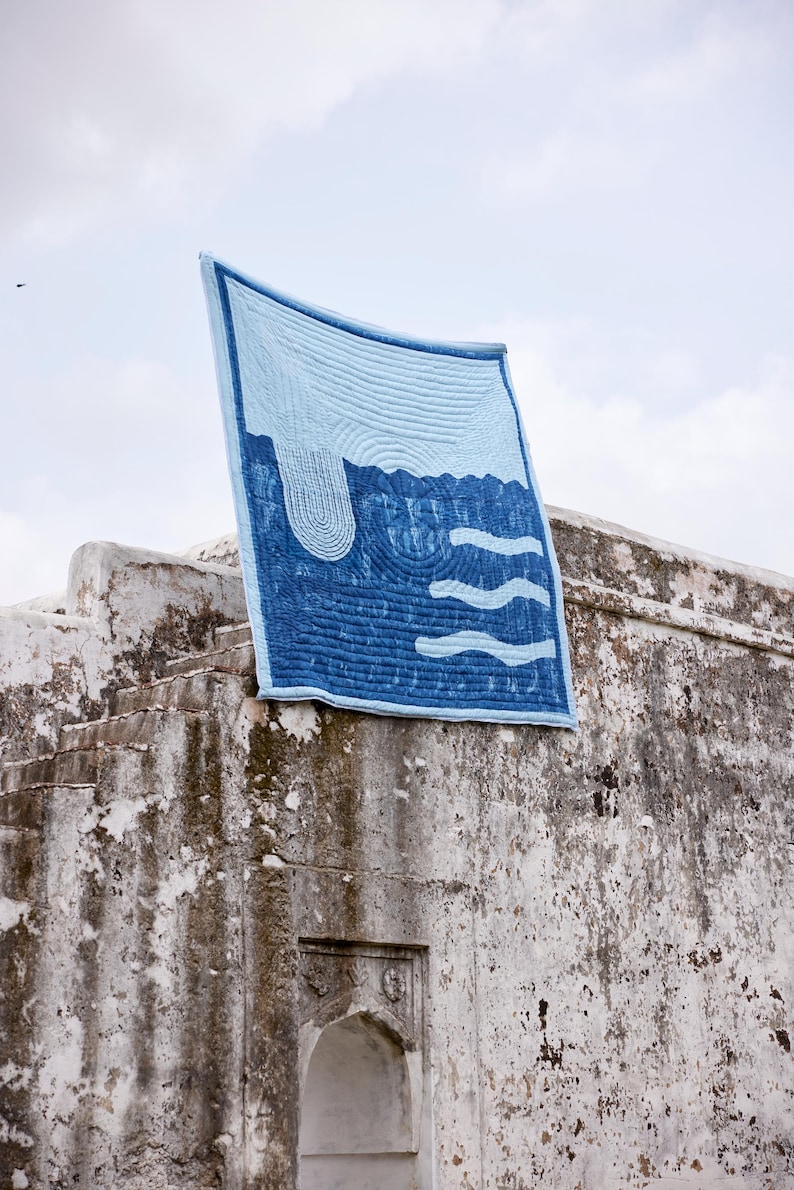 K&ouml;nnte beinhalten: Ein blau-wei&szlig;es Quilt mit einem vom Wasser inspirierten Design. Das Quilt hat einen hellblauen Hintergrund mit dunkelblauen Wellen- und Wasserfallmustern. Das Quilt wird im Freien &uuml;ber einer verwitterten Steinstruktur drapiert.