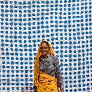 May include: A woman wearing a yellow head covering and matching skirt, standing in front of a white and blue star-patterned textile. The woman is also wearing a gray and black checkered shirt.
