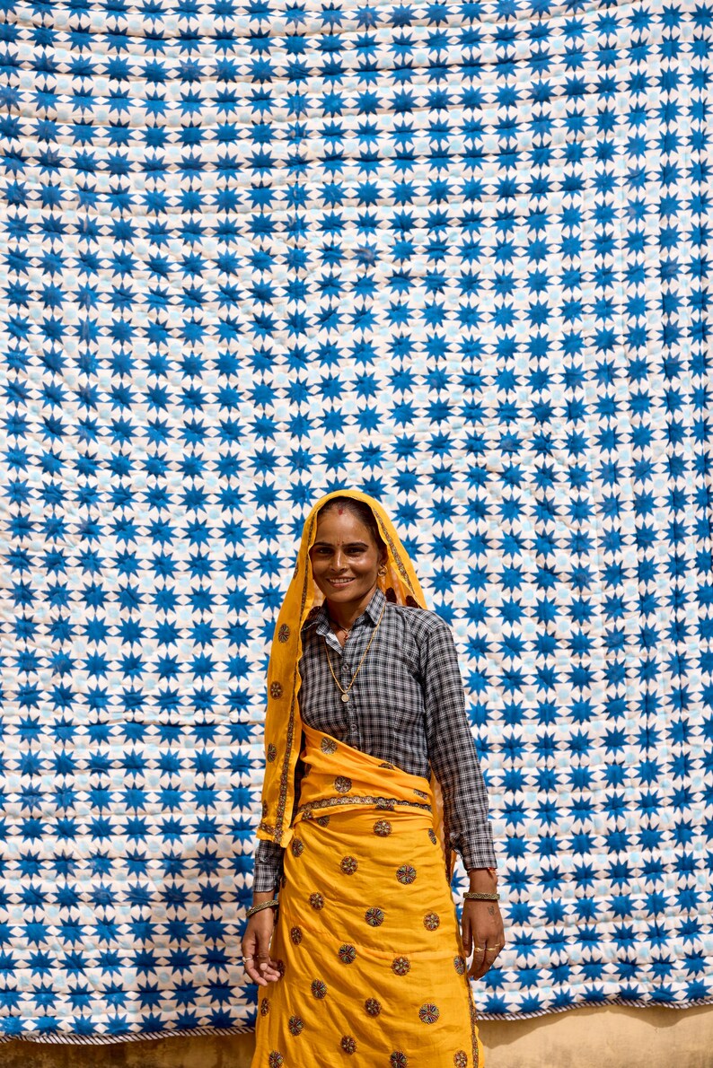 May include: A woman wearing a yellow head covering and matching skirt, standing in front of a white and blue star-patterned textile. The woman is also wearing a gray and black checkered shirt.