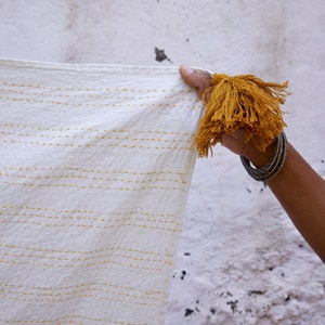 Peut inclure: Un textile blanc orn&eacute; de rang&eacute;es horizontales de points de couture jaunes et d'un grand pompon d&eacute;coratif jaune moutarde fix&eacute; &agrave; un coin. Le tissu pr&eacute;sente une surface textur&eacute;e et est maintenu pour montrer les d&eacute;tails des points et du pompon.