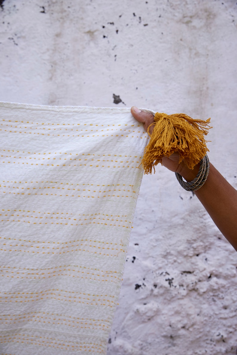 Peut inclure: Un textile blanc orn&eacute; de rang&eacute;es horizontales de points de couture jaunes et d'un grand pompon d&eacute;coratif jaune moutarde fix&eacute; &agrave; un coin. Le tissu pr&eacute;sente une surface textur&eacute;e et est maintenu pour montrer les d&eacute;tails des points et du pompon.