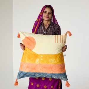 May include: A rectangular decorative pillow with a cream-colored background and orange, yellow, and blue geometric patterns. The pillow has orange tassels on each corner. The pillow is held by a person wearing a purple head covering and dress.