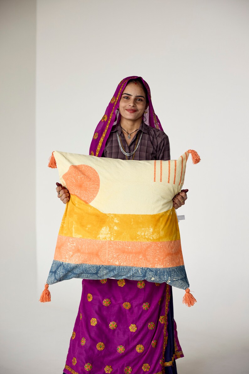 May include: A rectangular decorative pillow with a cream-colored background and orange, yellow, and blue geometric patterns. The pillow has orange tassels on each corner. The pillow is held by a person wearing a purple head covering and dress.