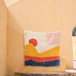 May include: A colorful, quilted wall hanging with a landscape design. The artwork features a red sun, yellow and orange mountains, and a blue base. The quilt is displayed on a light yellow wall, adding a touch of warmth to the space.