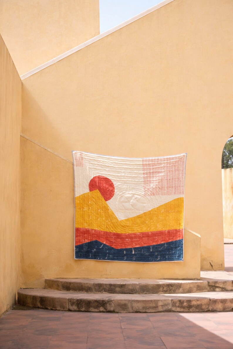 May include: A colorful, quilted wall hanging with a landscape design. The artwork features a red sun, yellow and orange mountains, and a blue base. The quilt is displayed on a light yellow wall, adding a touch of warmth to the space.
