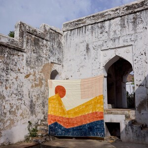 May include: A colorful quilt with a landscape design, featuring a large orange sun and layered mountain shapes in yellow, orange, and blue. The quilt is hanging against a weathered white wall with arched doorways.