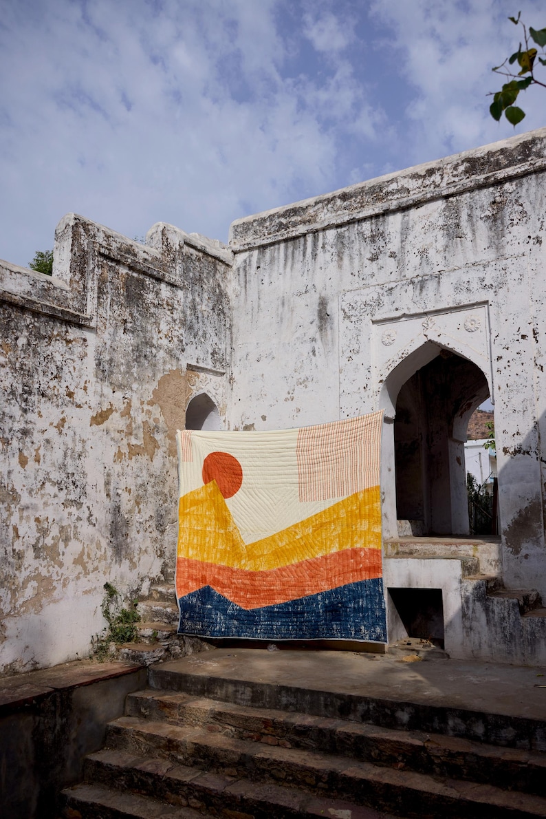 May include: A colorful quilt with a landscape design, featuring a large orange sun and layered mountain shapes in yellow, orange, and blue. The quilt is hanging against a weathered white wall with arched doorways.