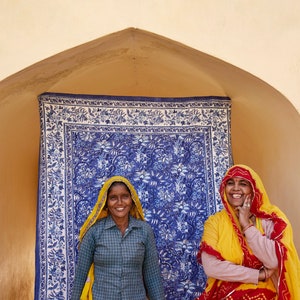 Puede incluir: Dos mujeres con saris coloridos se paran frente a un tel&oacute;n de fondo de tela con estampado azul y blanco. La mujer de la izquierda lleva un sari amarillo y verde con una camisa a cuadros. La mujer de la derecha lleva un sari amarillo con detalles rojos y blancos.