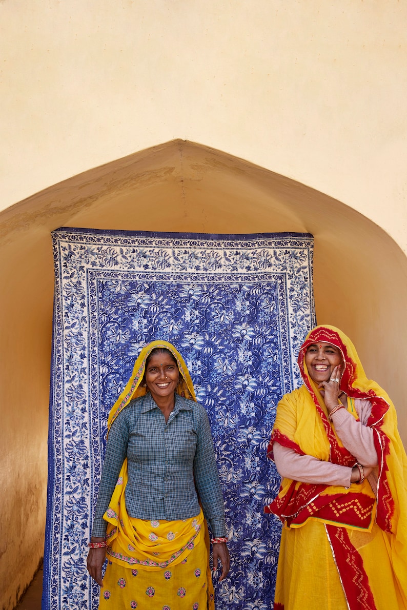 Puede incluir: Dos mujeres con saris coloridos se paran frente a un tel&oacute;n de fondo de tela con estampado azul y blanco. La mujer de la izquierda lleva un sari amarillo y verde con una camisa a cuadros. La mujer de la derecha lleva un sari amarillo con detalles rojos y blancos.