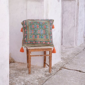 Pu&ograve; includere: Un cuscino con motivo colorato e nappe arancioni si trova su uno sgabello di legno contro un muro bianco.