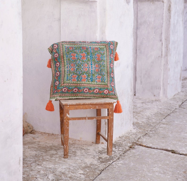Pu&ograve; includere: Un cuscino con motivo colorato e nappe arancioni si trova su uno sgabello di legno contro un muro bianco.