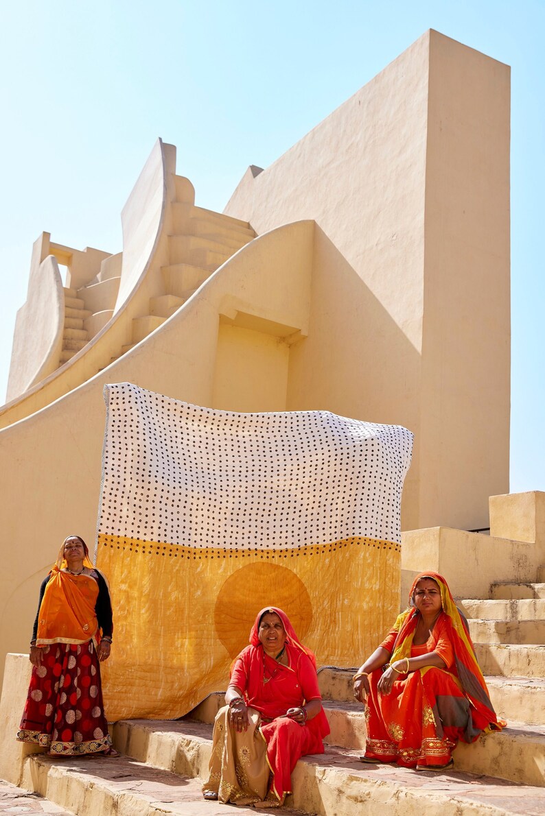 May include: Three women wearing colorful saris stand in front of a large, yellow building with a patterned fabric draped over a wall. The fabric is white with black polka dots and a yellow sun design.