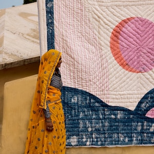 May include: A large, colorful quilt with a geometric design featuring a pink circle and blue accents. A person wearing a yellow outfit with a matching head covering stands in front of the quilt.