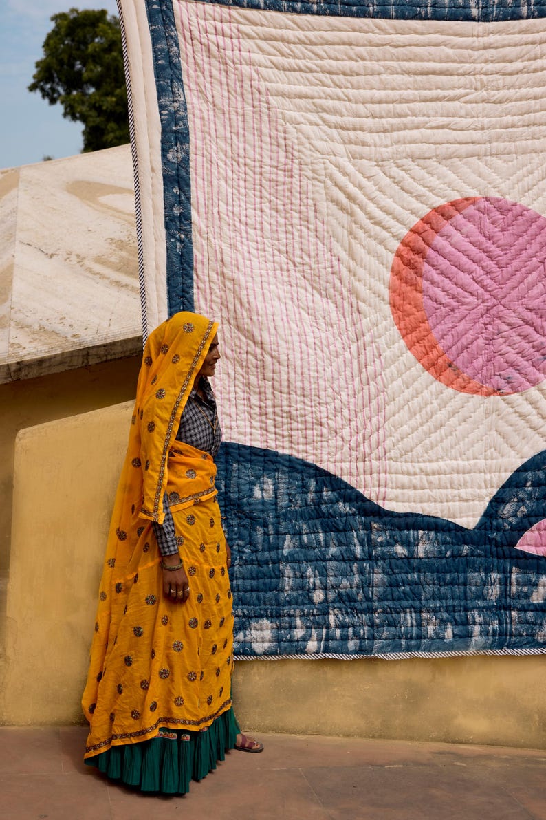 May include: A large, colorful quilt with a geometric design featuring a pink circle and blue accents. A person wearing a yellow outfit with a matching head covering stands in front of the quilt.