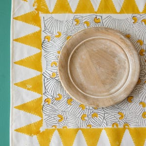 Puede incluir: Un mantel individual de algodón blanco con un patrón geométrico amarillo y negro. Un plato de madera se coloca encima del mantel individual.