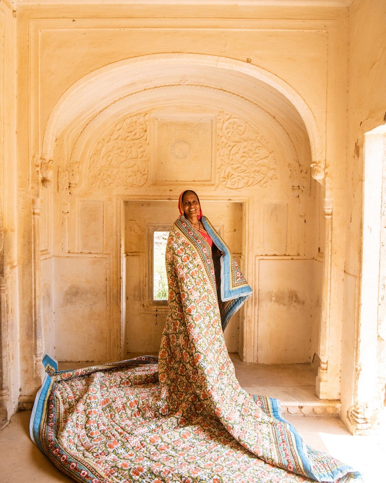 Puede incluir: Una mujer con un pa&ntilde;uelo rojo en la cabeza se encuentra en una puerta, sosteniendo una gran colcha con estampado floral. La colcha est&aacute; extendida en el suelo, creando un dise&ntilde;o colorido e intrincado.