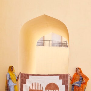 May include: Two women stand in front of a white and brown patterned rug with two large brown circles. The rug is hanging on a wall with a yellow archway behind it. The women are wearing traditional Indian clothing. One woman is wearing a yellow headscarf and a blue dress. The other woman is wearing an orange headscarf and a blue and orange dress.