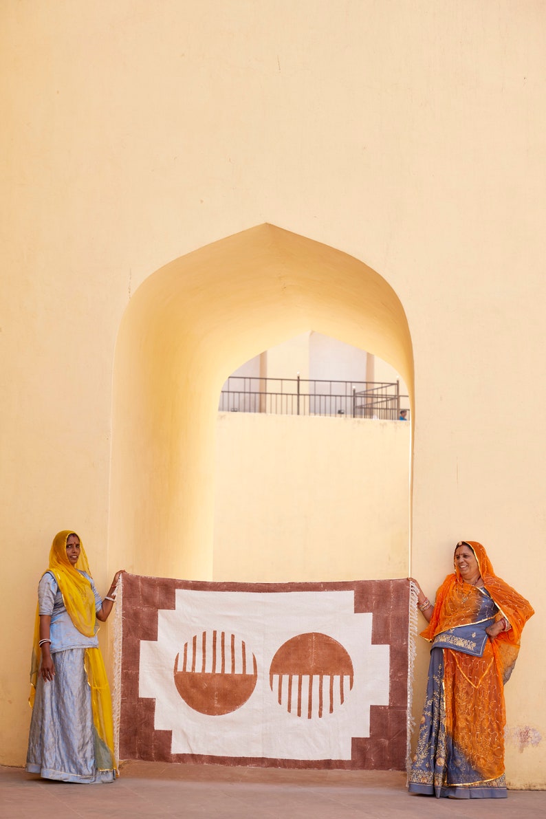 May include: Two women stand in front of a white and brown patterned rug with two large brown circles. The rug is hanging on a wall with a yellow archway behind it. The women are wearing traditional Indian clothing. One woman is wearing a yellow headscarf and a blue dress. The other woman is wearing an orange headscarf and a blue and orange dress.