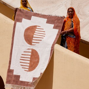 May include: Two women in traditional Indian clothing hold up a brown and white rug with a geometric pattern. The rug has two large, half-circle shapes in brown with white stripes.