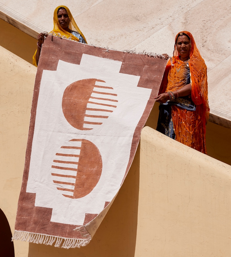 May include: Two women in traditional Indian clothing hold up a brown and white rug with a geometric pattern. The rug has two large, half-circle shapes in brown with white stripes.