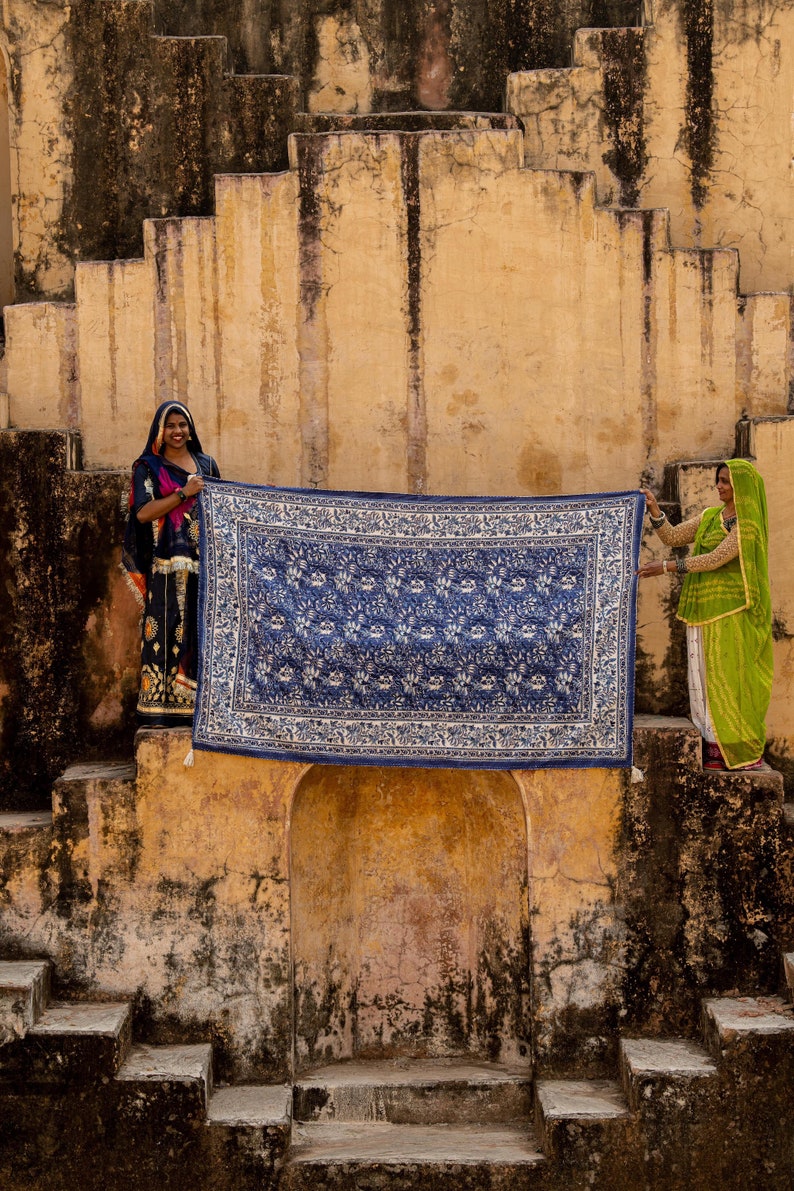 Puede incluir: Dos mujeres con ropa tradicional india se encuentran en un conjunto de escalones de piedra, sosteniendo una alfombra azul y blanca con estampado.