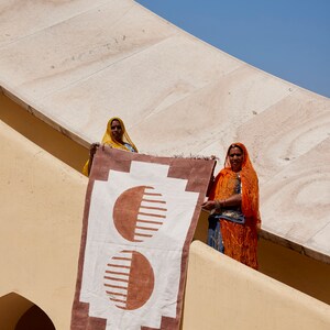 May include: Two women in colorful head scarves hold up a white and brown rug with a geometric pattern of two overlapping circles. The rug has a fringe border.