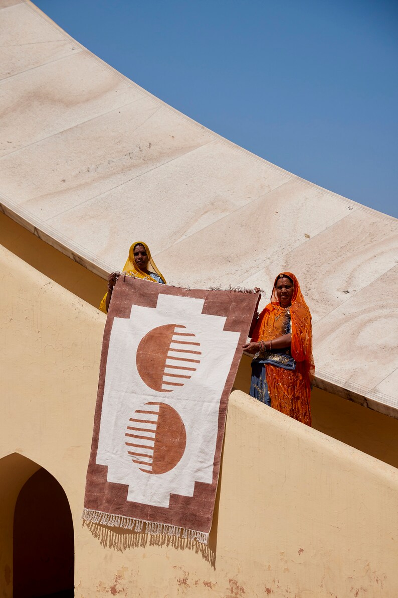 May include: Two women in colorful head scarves hold up a white and brown rug with a geometric pattern of two overlapping circles. The rug has a fringe border.