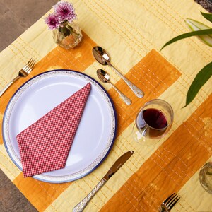 Puede incluir: Un plato blanco con un borde azul y una servilleta a cuadros rojos y blancos se encuentra sobre un mantel amarillo y naranja. Hay dos tenedores dorados y dos cucharas de plata en la mesa, junto con una copa de vino tinto.