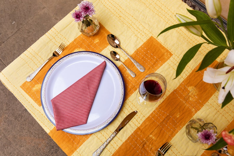 Puede incluir: Un plato blanco con un borde azul y una servilleta a cuadros rojos y blancos se encuentra sobre un mantel amarillo y naranja. Hay dos tenedores dorados y dos cucharas de plata en la mesa, junto con una copa de vino tinto.