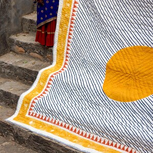 May include: A white quilt with navy blue diagonal stripes and a large yellow circle. The quilt has a yellow border with orange triangle accents. The quilt is folded over a stone staircase.