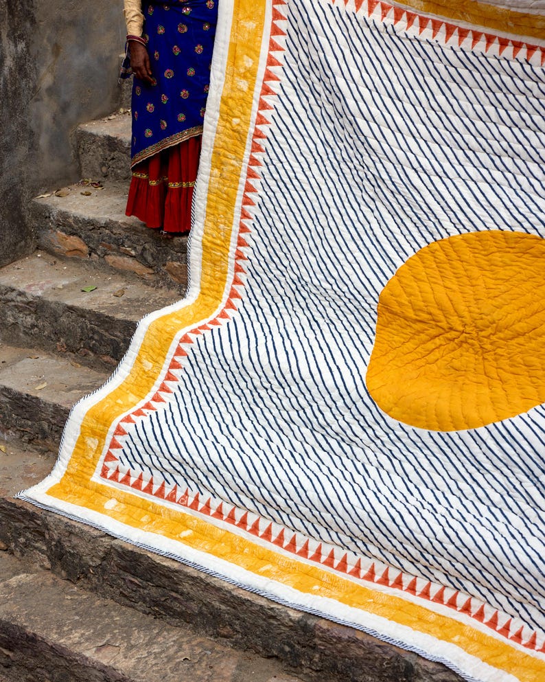 May include: A white quilt with navy blue diagonal stripes and a large yellow circle. The quilt has a yellow border with orange triangle accents. The quilt is folded over a stone staircase.