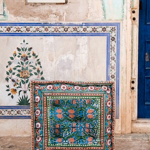 May include: A blue and green patterned floor cushion with a floral design. The cushion is sitting in front of a wall with a blue and white tiled design.