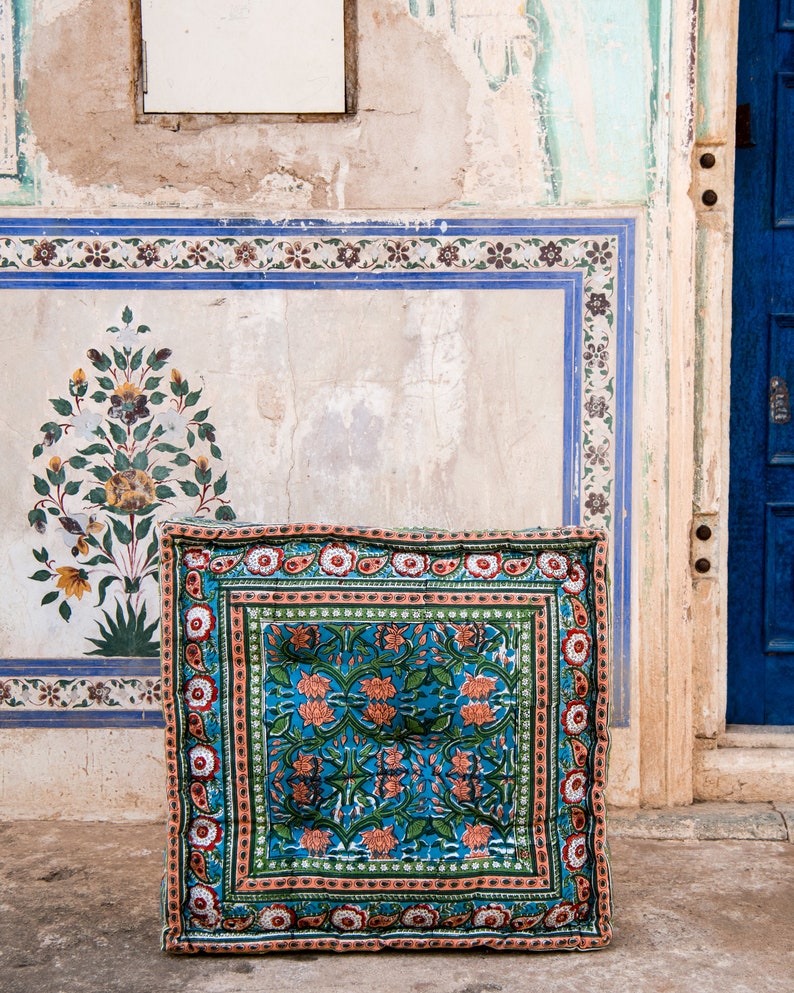 May include: A blue and green patterned floor cushion with a floral design. The cushion is sitting in front of a wall with a blue and white tiled design.