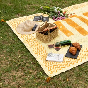 Puede incluir: Una manta de picnic amarilla y naranja con un patr&oacute;n de chevron, una cesta de mimbre, dos copas de vino, una botella de vino, dos croissants, un libro, un sombrero de paja y gafas de sol.