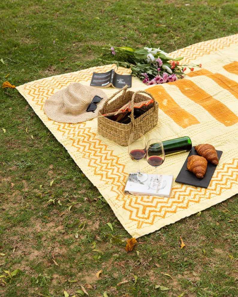 Puede incluir: Una manta de picnic amarilla y naranja con un patr&oacute;n de chevron, una cesta de mimbre, dos copas de vino, una botella de vino, dos croissants, un libro, un sombrero de paja y gafas de sol.