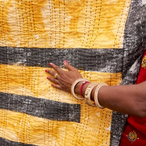 May include: A hand with red nail polish rests on a yellow and black striped fabric. The fabric has a quilted texture and is decorated with small black stitches. The hand is wearing several gold bangles and a red string bracelet.